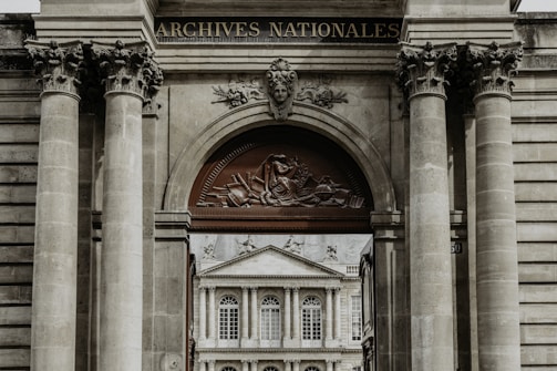 A grand architectural structure with ornate columns and a decorative archway, featuring a relief sculpture and the words 'Archives Nationales'. Through the archway, there is a view of a classical-style building with large windows and ornate detailing.