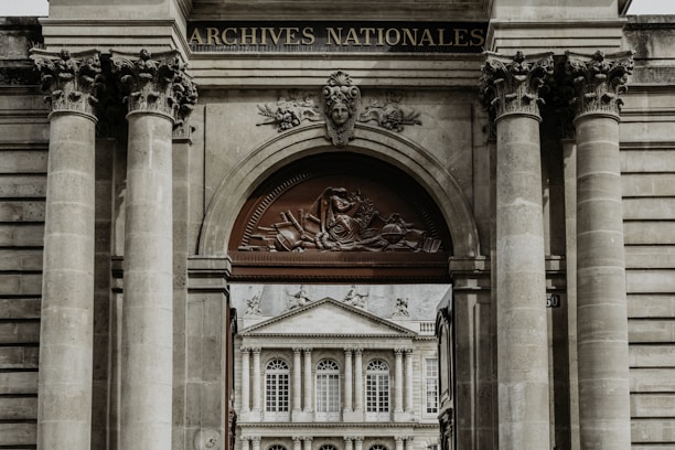 A grand architectural structure with ornate columns and a decorative archway, featuring a relief sculpture and the words 'Archives Nationales'. Through the archway, there is a view of a classical-style building with large windows and ornate detailing.