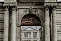 A grand architectural structure with ornate columns and a decorative archway, featuring a relief sculpture and the words 'Archives Nationales'. Through the archway, there is a view of a classical-style building with large windows and ornate detailing.