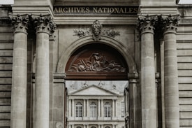 A grand architectural structure with ornate columns and a decorative archway, featuring a relief sculpture and the words 'Archives Nationales'. Through the archway, there is a view of a classical-style building with large windows and ornate detailing.