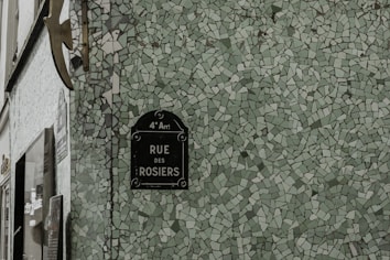 A mosaic-patterned wall with a street sign reading 'RUE DES ROSIERS' in the 4th district. The wall features small, irregular green tiles, creating an artistic effect.