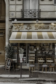 A quaint caf&eacute; storefront with vintage-style signage reading 'La Bossue', featuring a striped awning in shades of cream and brown. There are small round tables and wooden chairs arranged on the sidewalk, indicating outdoor seating. Potted plants and a chalkboard menu stand add to the charm of the setting. The building facade includes decorative ironwork and classic architecture, with a central window above the caf&eacute;.
