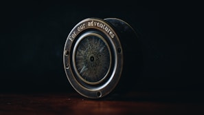 An antique-style compass with the phrase 'for our adventures' engraved on its brass casing sits on a wooden surface. The dim lighting highlights the intricate design on the compass face, creating a sense of mystery and nostalgia.