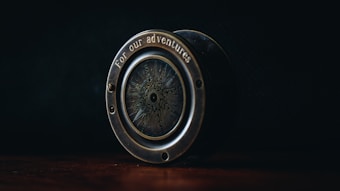 An antique-style compass with the phrase 'for our adventures' engraved on its brass casing sits on a wooden surface. The dim lighting highlights the intricate design on the compass face, creating a sense of mystery and nostalgia.