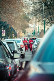 A traffic scene on a city street with several cars lined up closely. Two individuals wearing red uniforms stand between the cars, possibly signaling or directing traffic. Leafless trees line the street, indicating a winter or late fall setting.