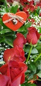 A bouquet of vibrant red roses surrounded by lush green leaves and delicate white baby's breath flowers. A heart-shaped decoration with a small silver heart and the word 'LOVE' adds a romantic touch to the arrangement.