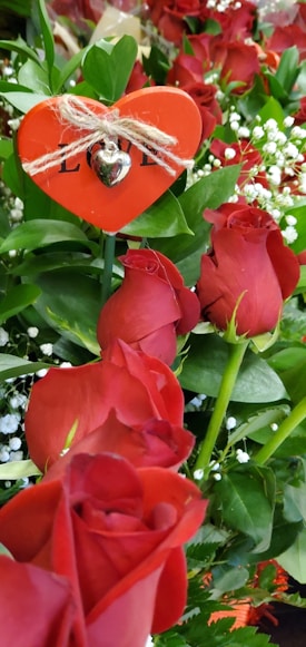 A bouquet of vibrant red roses surrounded by lush green leaves and delicate white baby's breath flowers. A heart-shaped decoration with a small silver heart and the word 'LOVE' adds a romantic touch to the arrangement.