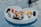 A heart-shaped wicker basket is floating on a pool, filled with a variety of breakfast items including glasses of orange juice, red juice, fruit slices, pastries, and yogurt parfaits. Silver carafes and neatly folded black napkins are also present, creating an elegant and luxurious setting.