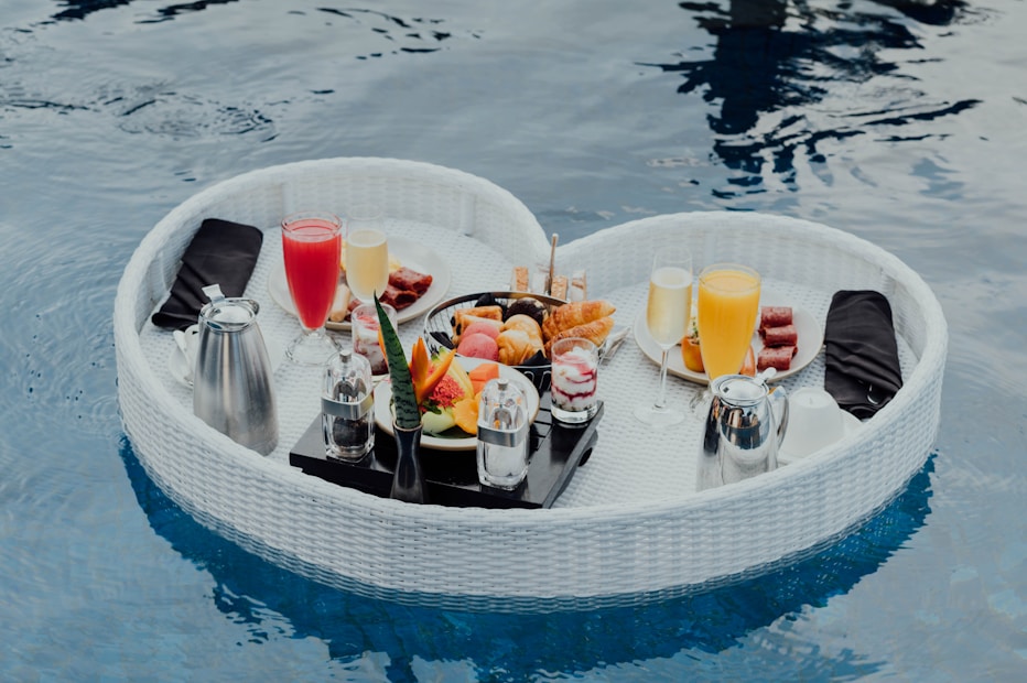 A heart-shaped wicker basket is floating on a pool, filled with a variety of breakfast items including glasses of orange juice, red juice, fruit slices, pastries, and yogurt parfaits. Silver carafes and neatly folded black napkins are also present, creating an elegant and luxurious setting.