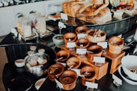 An assortment of wooden containers filled with various dried foods and spices is arranged on a countertop. In the background, glass jars filled with different dry goods are visible, alongside a decorative statue. The setup appears to be organized, possibly for a buffet or self-service station.