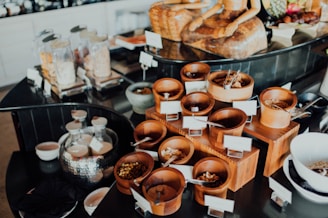 An assortment of wooden containers filled with various dried foods and spices is arranged on a countertop. In the background, glass jars filled with different dry goods are visible, alongside a decorative statue. The setup appears to be organized, possibly for a buffet or self-service station.