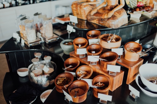 An assortment of wooden containers filled with various dried foods and spices is arranged on a countertop. In the background, glass jars filled with different dry goods are visible, alongside a decorative statue. The setup appears to be organized, possibly for a buffet or self-service station.