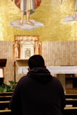A person is kneeling in prayer inside a church, facing an altar adorned with a golden mosaic and religious symbols. The person is wearing a dark hoodie and sits in a pew, surrounded by wooden furniture and small green plants. The setting conveys a sense of tranquility and spirituality.