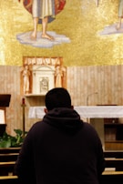 A person is kneeling in prayer inside a church, facing an altar adorned with a golden mosaic and religious symbols. The person is wearing a dark hoodie and sits in a pew, surrounded by wooden furniture and small green plants. The setting conveys a sense of tranquility and spirituality.