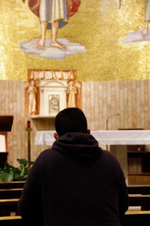 A person is kneeling in prayer inside a church, facing an altar adorned with a golden mosaic and religious symbols. The person is wearing a dark hoodie and sits in a pew, surrounded by wooden furniture and small green plants. The setting conveys a sense of tranquility and spirituality.