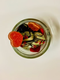 An overhead view of a glass jar filled with assorted candies, including heart-shaped gummies and chocolates drizzled with stripes and sprinkles. The heart-shaped gummy sits prominently on the jar's rim, adding a vibrant pop of red against the sweet contents inside.