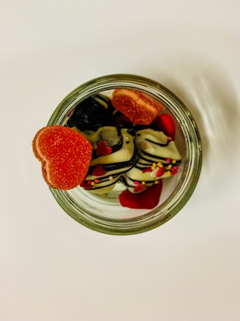 An overhead view of a glass jar filled with assorted candies, including heart-shaped gummies and chocolates drizzled with stripes and sprinkles. The heart-shaped gummy sits prominently on the jar's rim, adding a vibrant pop of red against the sweet contents inside.