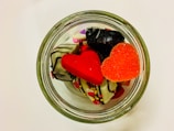 A glass jar is filled with colorful, heart-shaped candies and chocolates decorated with icing and sprinkles. The candies are vibrant with red and orange hues, complementing the white chocolate pieces.