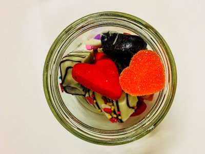 A glass jar is filled with colorful, heart-shaped candies and chocolates decorated with icing and sprinkles. The candies are vibrant with red and orange hues, complementing the white chocolate pieces.