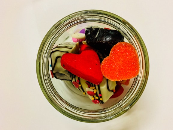 A glass jar is filled with colorful, heart-shaped candies and chocolates decorated with icing and sprinkles. The candies are vibrant with red and orange hues, complementing the white chocolate pieces.