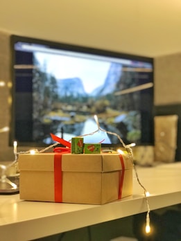 A gift box wrapped with a red ribbon is placed on a table. The box is adorned with fairy lights and has two green candy packages on top. In the background, a computer monitor displays a scenic landscape featuring mountains and trees.