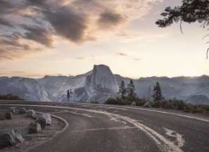 gray asphalt road near green trees and mountain under cloudy sky during daytime