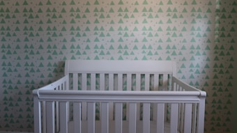 A white crib is positioned against a wall covered in a pattern of teal and white triangles. The crib has a simple, classic design with slatted sides and a high backrest.