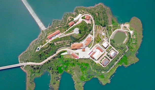 An aerial view of an island surrounded by water, featuring a complex with multiple buildings and landscaped areas. The island is connected to the mainland by a bridge, and it includes pathways, vegetation, and park-like settings.