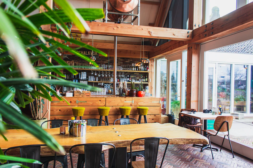 The image depicts a cozy restaurant interior with wooden beams and walls, creating an inviting atmosphere. The space is filled with natural light from large windows that offer a view of trees outside. In the center of the room stands a long wooden bar with green stools on either side, suggesting a place for patrons to enjoy their meals or drinks.

The restaurant features a dining area with several tables and chairs arranged around them, as well as a kitchenette equipped with a sink and countertop. The walls are adorned with various bottles of wine displayed in the background, indicating that the establishment offers a selection of beverages. A potted plant is also present on one side of the room, adding to the overall ambiance.