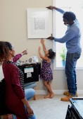 A family is engaging in a home activity, where a man, a woman, and a child are involved in hanging a framed alphabet print on the wall. The woman is kneeling and pointing at the artwork while the child is reaching upwards. The setting is a living room with moving boxes labeled 'Hive Boxx', indicating they might be unpacking or decorating a new home.