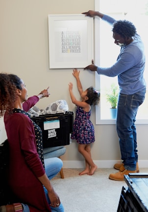 A family is engaging in a home activity, where a man, a woman, and a child are involved in hanging a framed alphabet print on the wall. The woman is kneeling and pointing at the artwork while the child is reaching upwards. The setting is a living room with moving boxes labeled 'Hive Boxx', indicating they might be unpacking or decorating a new home.