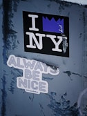 A textured wall features two distinct stickers. One sticker displays the phrase 'I NY' with a purple crown symbol replacing the heart. Below it, another sticker reads 'ALWAYS BE NICE' in bold letters accompanied by the hashtag #AlwaysBeNice.