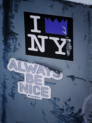 A textured wall features two distinct stickers. One sticker displays the phrase 'I NY' with a purple crown symbol replacing the heart. Below it, another sticker reads 'ALWAYS BE NICE' in bold letters accompanied by the hashtag #AlwaysBeNice.