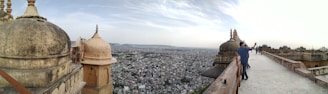 A panoramic view of a historical cityscape with ancient architectural structures featuring domes and intricate designs. A pathway leads along the edge of the buildings, with people walking and taking photos. The expansive city stretches into the distance under a partly cloudy sky.