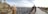 A panoramic view of a historical cityscape with ancient architectural structures featuring domes and intricate designs. A pathway leads along the edge of the buildings, with people walking and taking photos. The expansive city stretches into the distance under a partly cloudy sky.