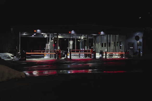 A dimly lit security checkpoint with barriers and multiple red lights reflecting on the wet ground. The area is mostly surrounded by darkness, with snow visible on the sides. Several overhead lights illuminate the structure, and there are signs present.