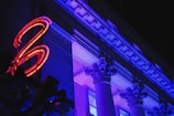 An architectural facade of a classical building lit with vibrant blue and purple lighting. The structure features ornate columns and intricate detailing. A red neon light in the shape of a number is visible on the left side, adding a modern contrast to the historical architecture.