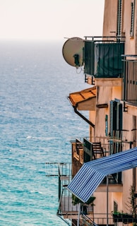 A building with several balconies overlooks a blue ocean. The balconies are equipped with metal railings and satellite dishes, and some have striped awnings and potted plants.