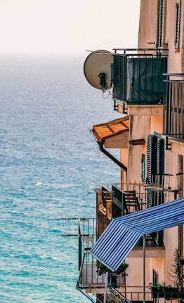 A building with several balconies overlooks a blue ocean. The balconies are equipped with metal railings and satellite dishes, and some have striped awnings and potted plants.