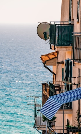 A building with several balconies overlooks a blue ocean. The balconies are equipped with metal railings and satellite dishes, and some have striped awnings and potted plants.