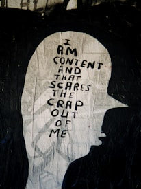 A piece of street art depicts the silhouette of a person wearing a cap, filled with a handwritten message. The words are bold and black against a textured white background, reading 'I am content and that scares the crap out of me.' The artwork is striking and thought-provoking, playing on contrasting themes of contentment and fear.