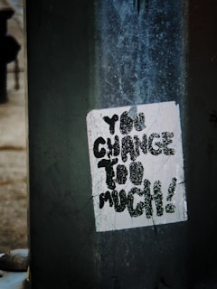 A close-up view of a weathered sticker on a dark, textured surface. The sticker contains the text 'You change too much!' in bold, black letters. The background has a gritty, urban feel with visible scratches and marks.