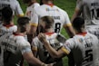 A group of rugby players wearing white jerseys with red and yellow accents stand together on the field. Some players have their arms around each other, suggesting team unity or celebration. The focus is on the back of their jerseys, displaying names and numbers.