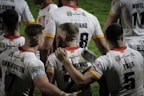 A group of rugby players wearing white jerseys with red and yellow accents stand together on the field. Some players have their arms around each other, suggesting team unity or celebration. The focus is on the back of their jerseys, displaying names and numbers.