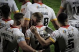A group of rugby players wearing white jerseys with red and yellow accents stand together on the field. Some players have their arms around each other, suggesting team unity or celebration. The focus is on the back of their jerseys, displaying names and numbers.