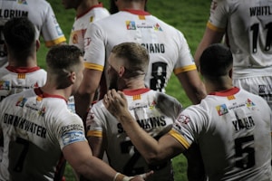 A group of rugby players wearing white jerseys with red and yellow accents stand together on the field. Some players have their arms around each other, suggesting team unity or celebration. The focus is on the back of their jerseys, displaying names and numbers.