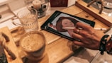 A hand is using a stylus to draw on a tablet, featuring an illustration of a woman's face. The scene is set on a wooden surface with a tray. Nearby, there are two glasses, one containing a hot beverage like coffee and the other filled with water. A candle and a decorative jar with pebbles are also visible in the background.