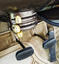 The image displays an old-fashioned car climate control system with dials labeled ‘DE-FOG,’ ‘DE-ICE,’ ‘LOW,’ ‘MED,’ ‘HI,’ ‘OFF,’ 'WARMER,' and levers marked ‘DEF,’ ‘AIR,’ ‘TEMP.’ The setup is situated above a carpeted floor and beneath a pair of foot pedals, typical of a classic vehicle interior.