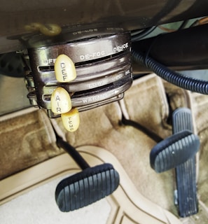 The image displays an old-fashioned car climate control system with dials labeled ‘DE-FOG,’ ‘DE-ICE,’ ‘LOW,’ ‘MED,’ ‘HI,’ ‘OFF,’ 'WARMER,' and levers marked ‘DEF,’ ‘AIR,’ ‘TEMP.’ The setup is situated above a carpeted floor and beneath a pair of foot pedals, typical of a classic vehicle interior.
