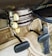 The image displays an old-fashioned car climate control system with dials labeled ‘DE-FOG,’ ‘DE-ICE,’ ‘LOW,’ ‘MED,’ ‘HI,’ ‘OFF,’ 'WARMER,' and levers marked ‘DEF,’ ‘AIR,’ ‘TEMP.’ The setup is situated above a carpeted floor and beneath a pair of foot pedals, typical of a classic vehicle interior.